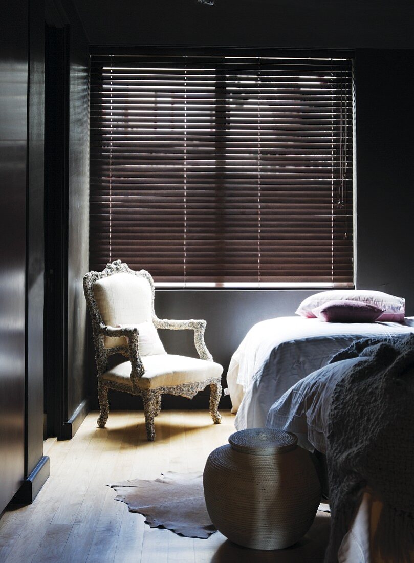 Postmodern armchair below window with closed louver blind in corner of bedroom