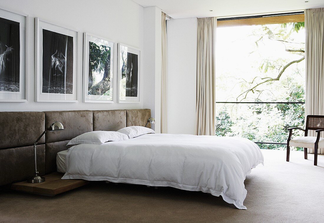 Doppelbett vor Wand mit halbhoch gepolsterten Lederpaneelen unter Fotogalerie, Blick in Garten durch raumhohes Fenster