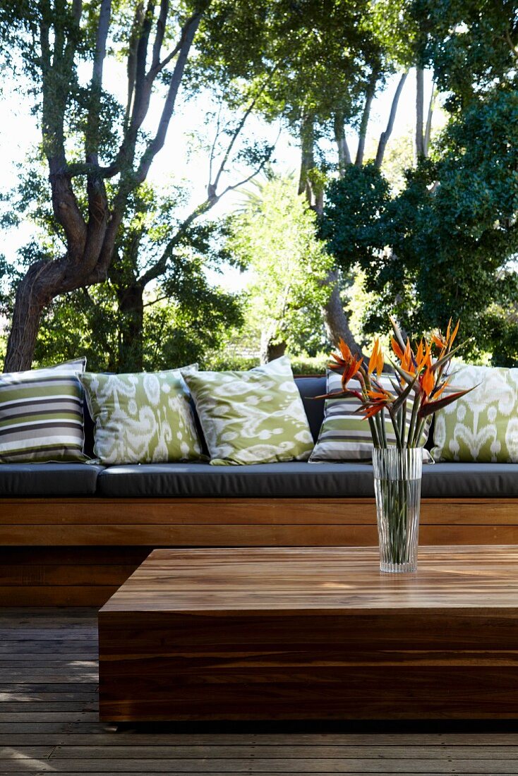 Vase mit Strelitzien auf Bodentisch aus tropischem Holz und Sitzbank mit Kissen auf Terrasse vor tropischem Garten