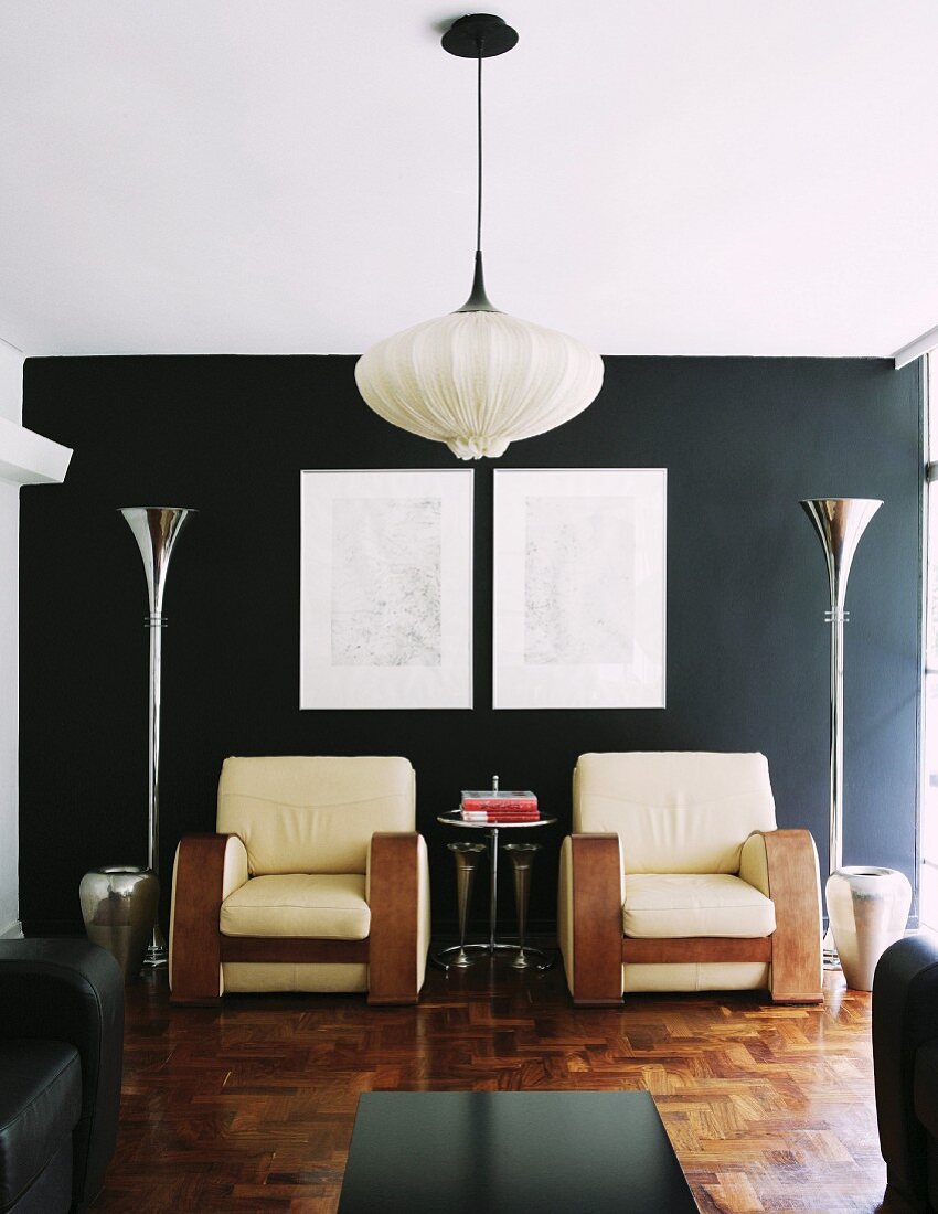 Hängeleuchte mit Stoffschirm in modernem Wohnzimmer mit mächtigen Sesseln vor schwarzer Wand zwischen trichterförmigen Designer-Stehleuchten