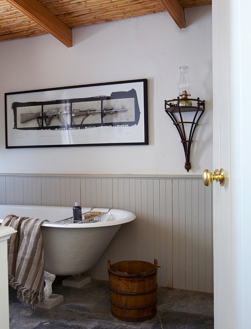 Wooden tub and free-standing bathtub against half-height, pale grey wooden panelling below framed picture on wall