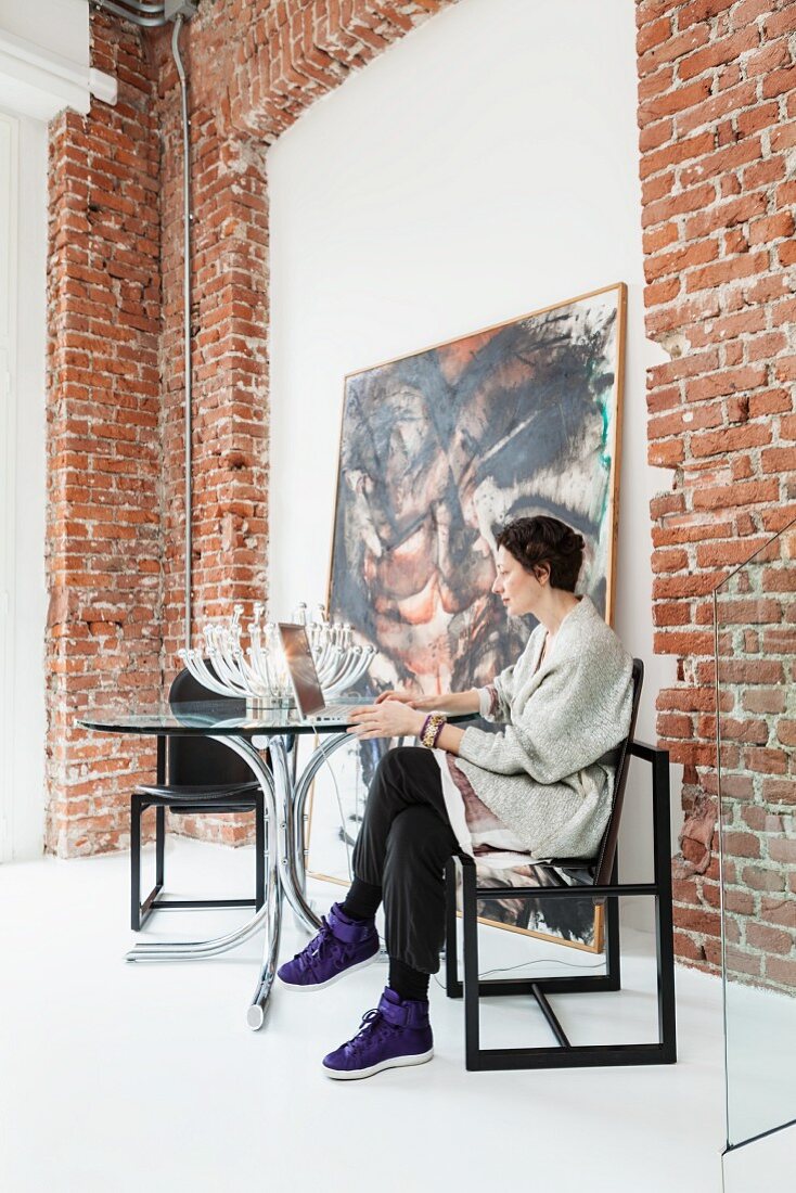 Frau auf Designerstuhl an rundem Glastisch vor an die Backsteinwand eines Loftraumes angelehntem Gemälde