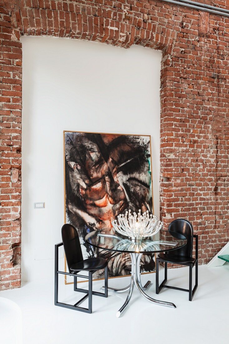 Blütenleuchte auf rundem Glastisch mit Designerstühlen vor einem abstrakten Gemälde in der Mauernische einer Backsteinwand