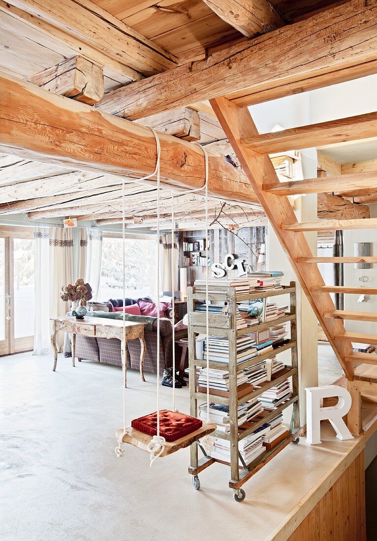 Child's swing hung from wooden beam next to bookshelves on castors in open-plan interior of wooden chalet