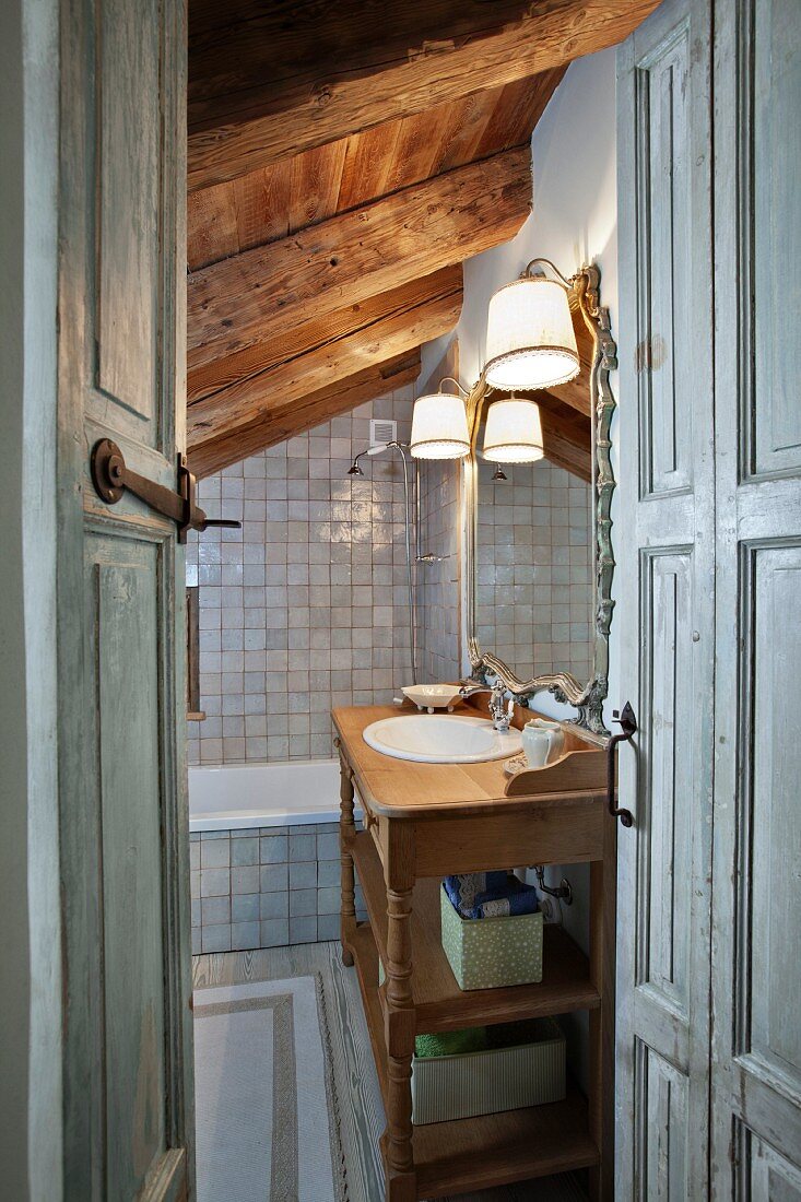 View into grey-tiled bathroom with rustic wooden washstand, mirror and sconce lamps