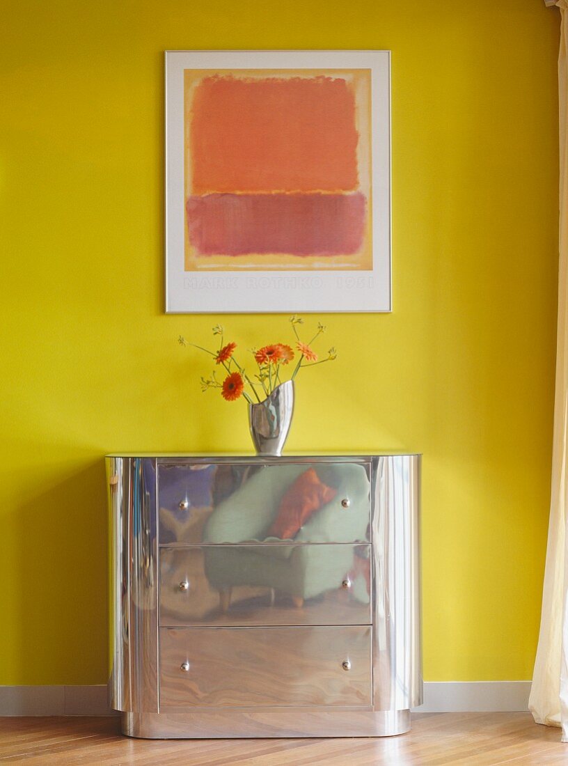 Blumenvase auf Metall-Kommode vor gelber Wand mit abstraktem Wandbild