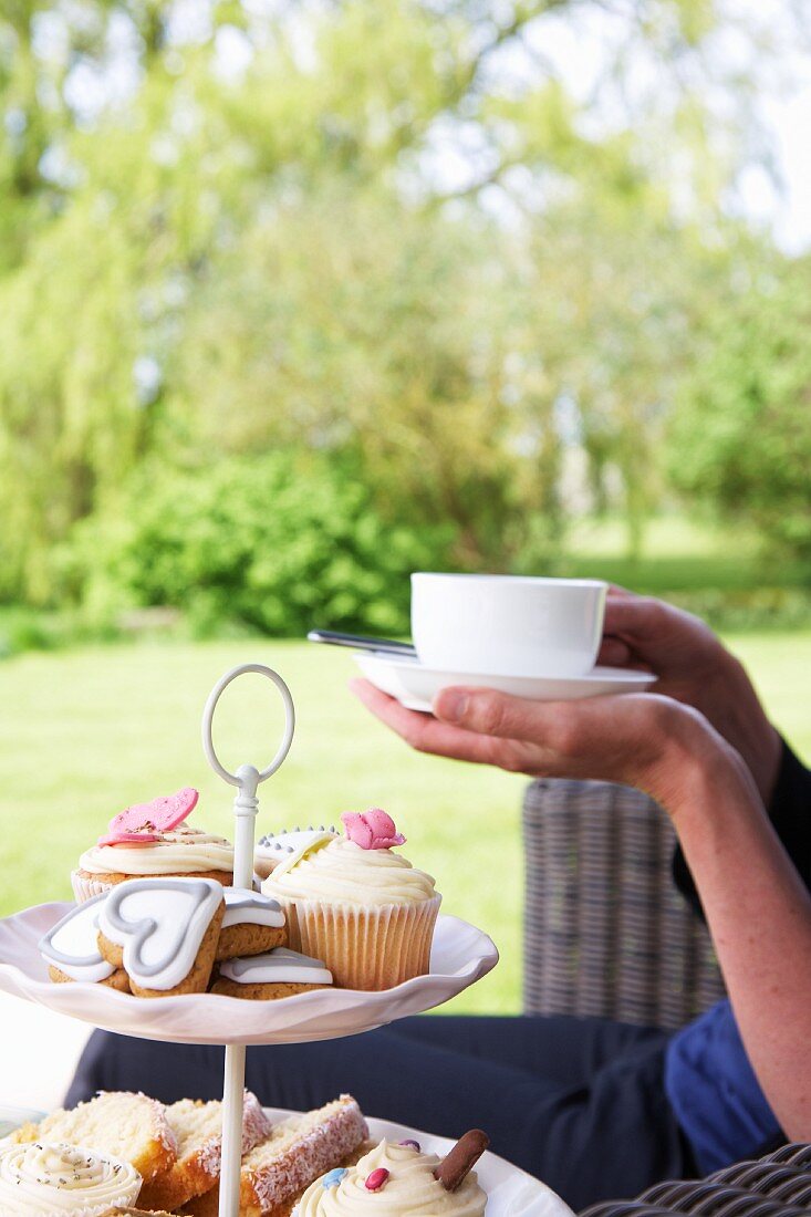 Frau mit Tasse Tee & Gebäck auf Etagere im Garten sitzend
