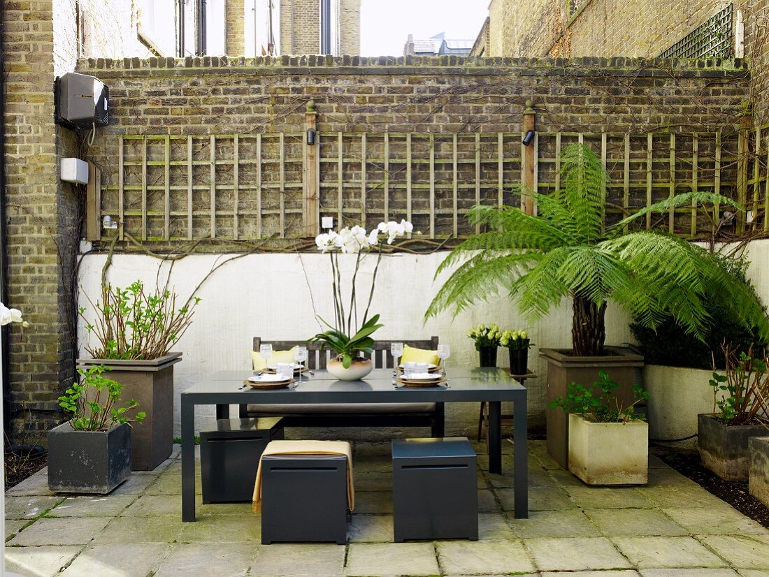 Sitzplatz in geschütztem Innenhof mit Backsteinmauern; elegante, graue Hocker an einem Tisch zwischen Pflanztöpfen