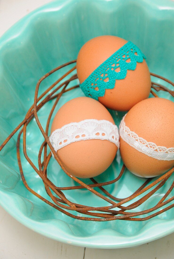Three Easter eggs with white and blue lace trim on nest of rusty, bent wire in pale blue bowl
