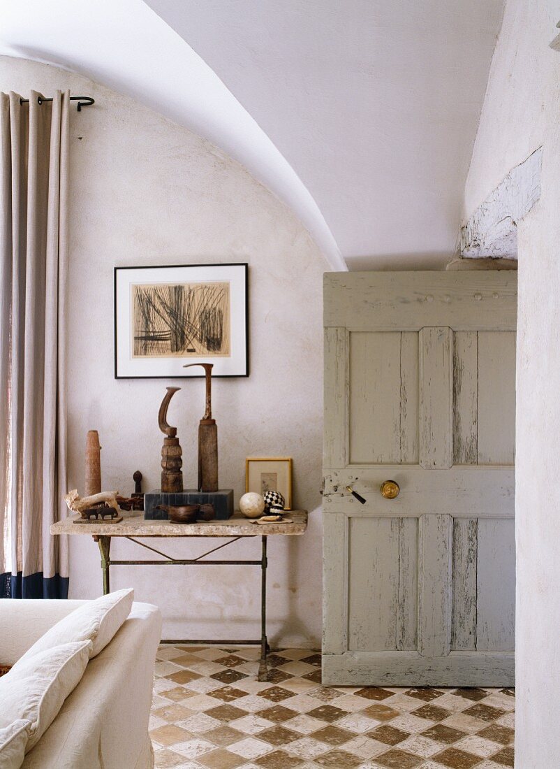 Kunstobjekte auf Beistelltisch und alte Holztür im Wohnraum eines historischen Gebäudes mit Kreuzgewölbe und Fliesenboden