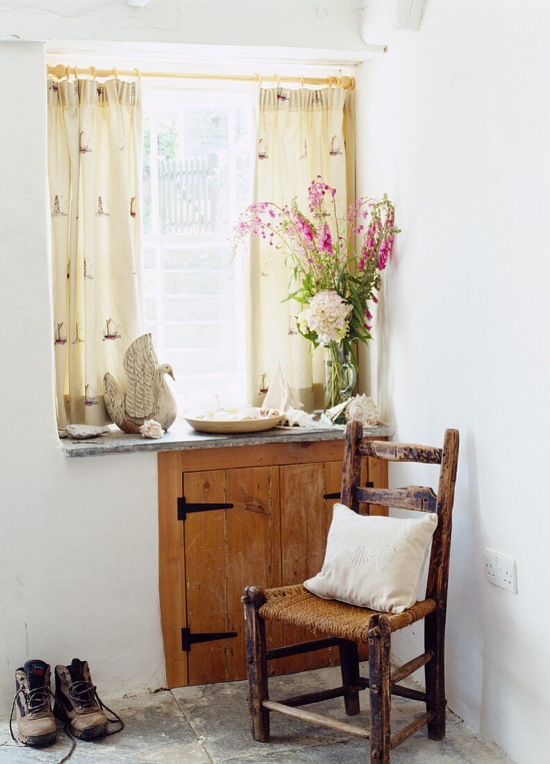 Praktischer Stauraum im Flur hinter Holztüren in der Brüstungswand eines Fensters; davor ein alter Geflechtstuhl und Wanderschuhe