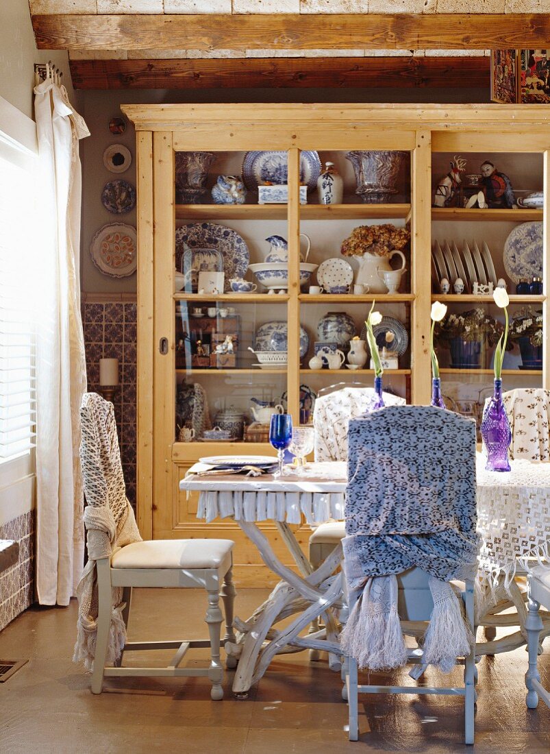 Mit Tüchern drapierte Holzstühle und Esstisch mit Beinen aus Astwerk vor einem Geschirrschrank mit Blauweiss-Porzellan