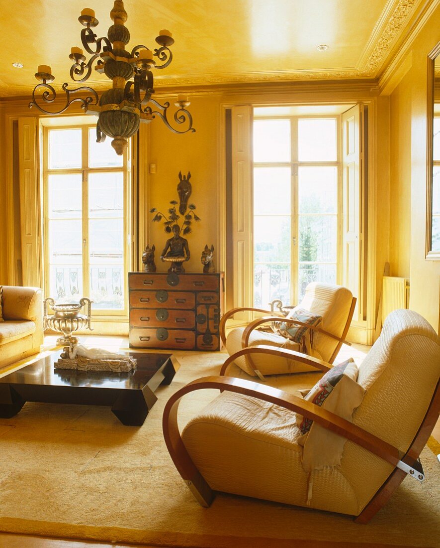 Gelb getönter Salon mit modernen Polstersesseln und Holztisch im dunklen Kolonialstil unter antikem Kronleuchter