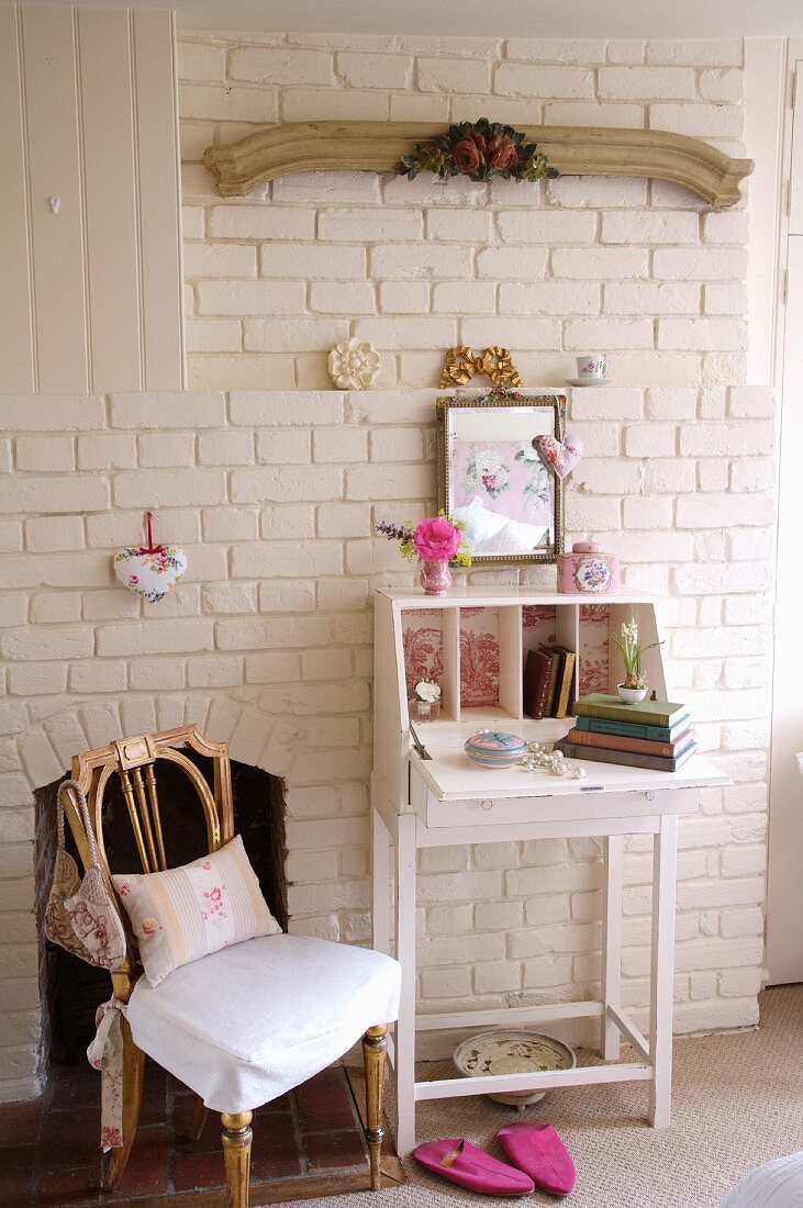 Holzstuhl neben geöffnetem Sekretär mit Bücherstapel und romantischen Nippes vor weiss geschlemmter Ziegelmauer