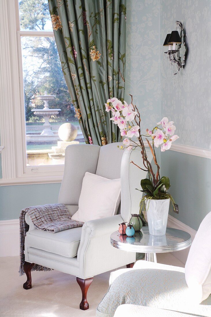 Orchidee auf Beistelltisch und eleganter Ohrensessel; Fenster zum Garten mit floral gemustertem Vorhang im Hintergrund