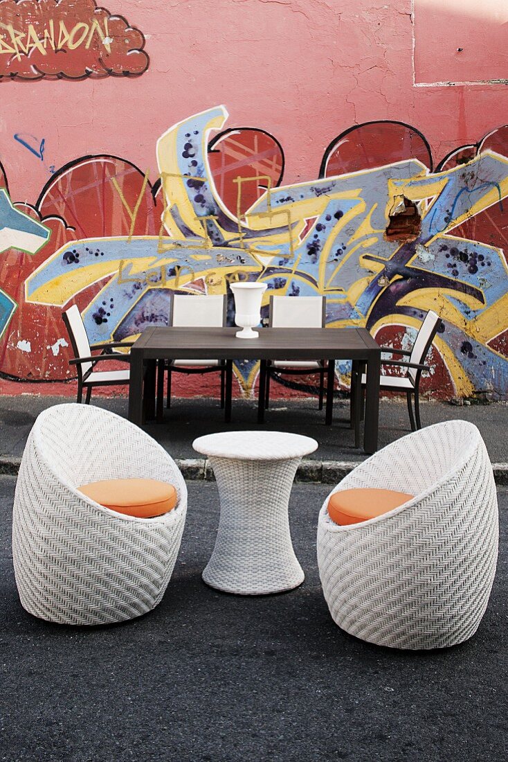 Gartentisch und Stühle sowie ein Tischchen aus Korbgeflecht mit zwei Schalenstühlen vor einer mit Graffiti bemalten Hauswand