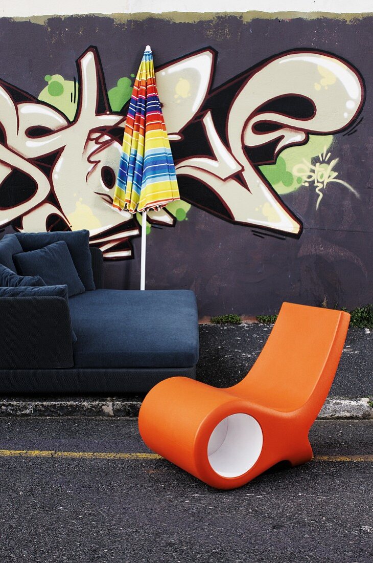 Sofa mit Sonnenschirm und orangefarbener Liegestuhl vor einer mit Graffiti bemalten Hauswand