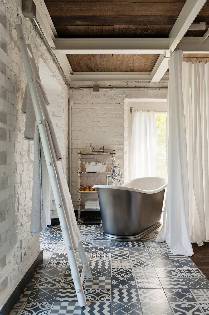 Leiter als Handtuchhalter, im Hintergrund freistehende Vintage Badewanne hinter Vorhang in Zimmerecke mit geweisselten Ziegelwänden und gemustertem Fliesenboden
