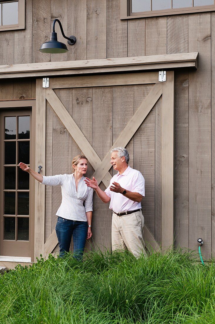 Mann und Frau unterhalten sich mit Gesten vor einem großen Schiebeladen einer Holzfassade in ländlichem Ambiente