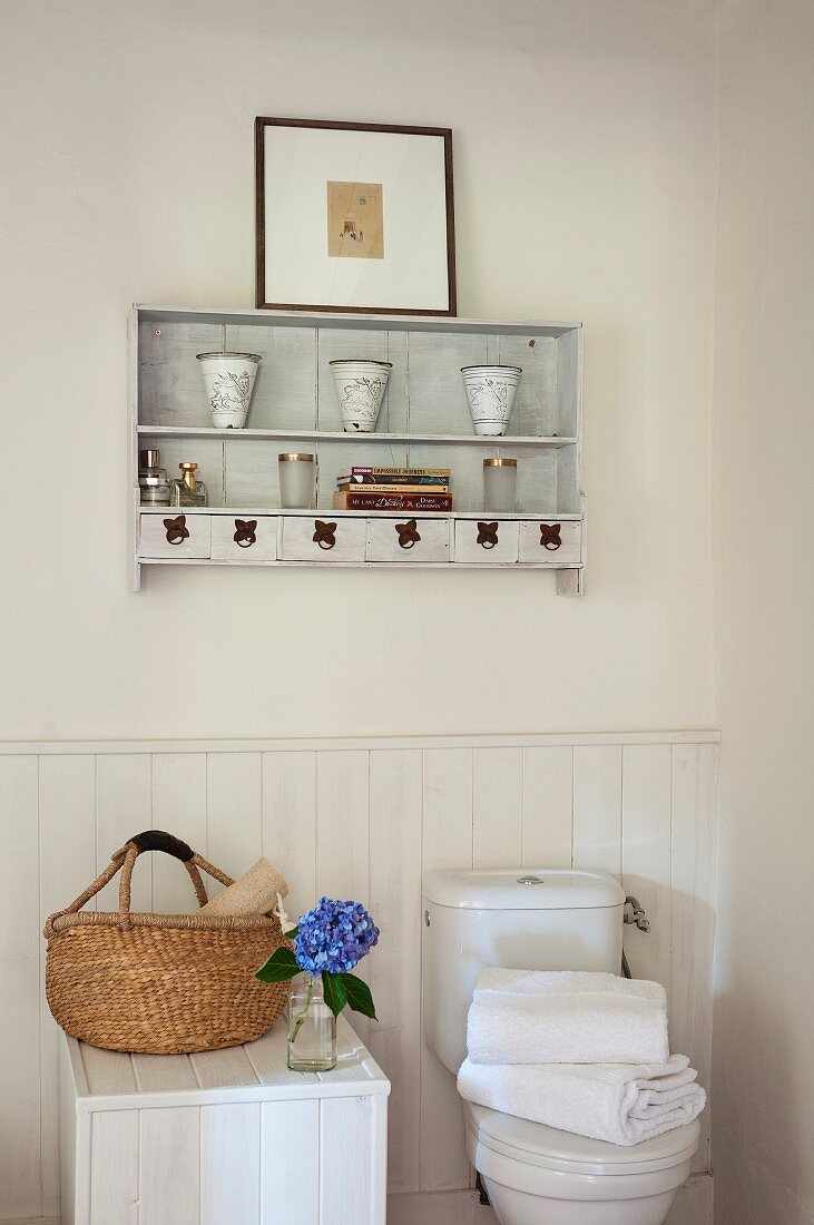 Basket on white, half-height wooden cabinet next to toilet and antique vases on wall-mounted shelves