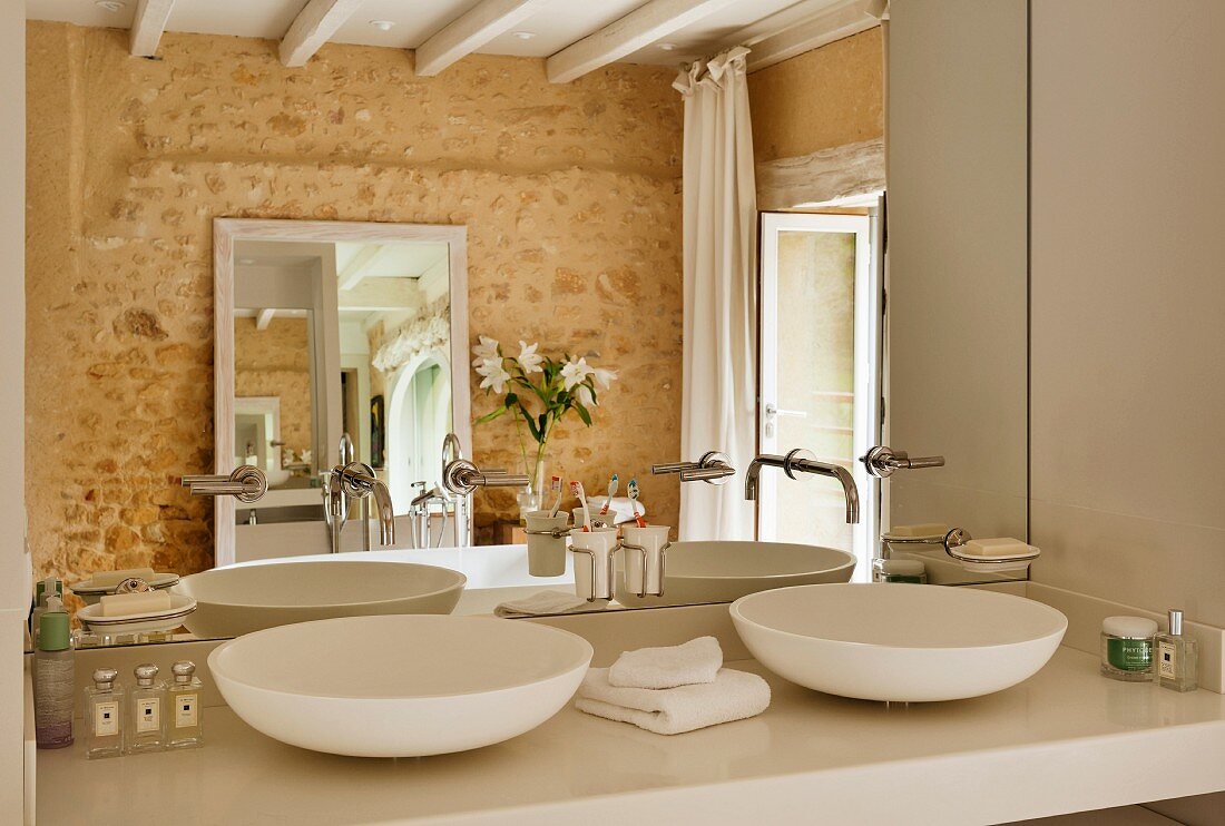 Twin countertop washbasins against mirrored wall in bathroom with stone wall