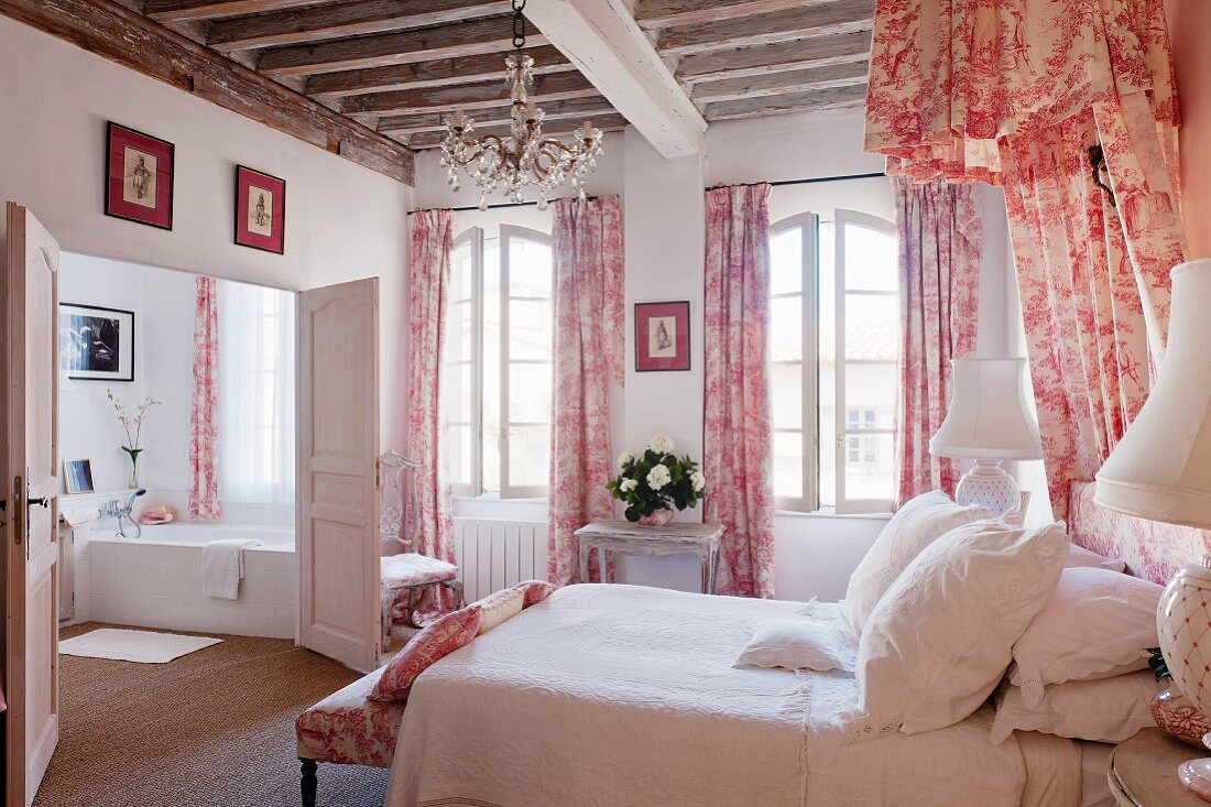 Romantisches Schlafzimmer mit rotweißem Toile-de-Jouy für Vorhänge und Betthimmel im Shabby-Chic Flair unter rustikaler Holzbalkendecke und Blick in Bad Ensuite