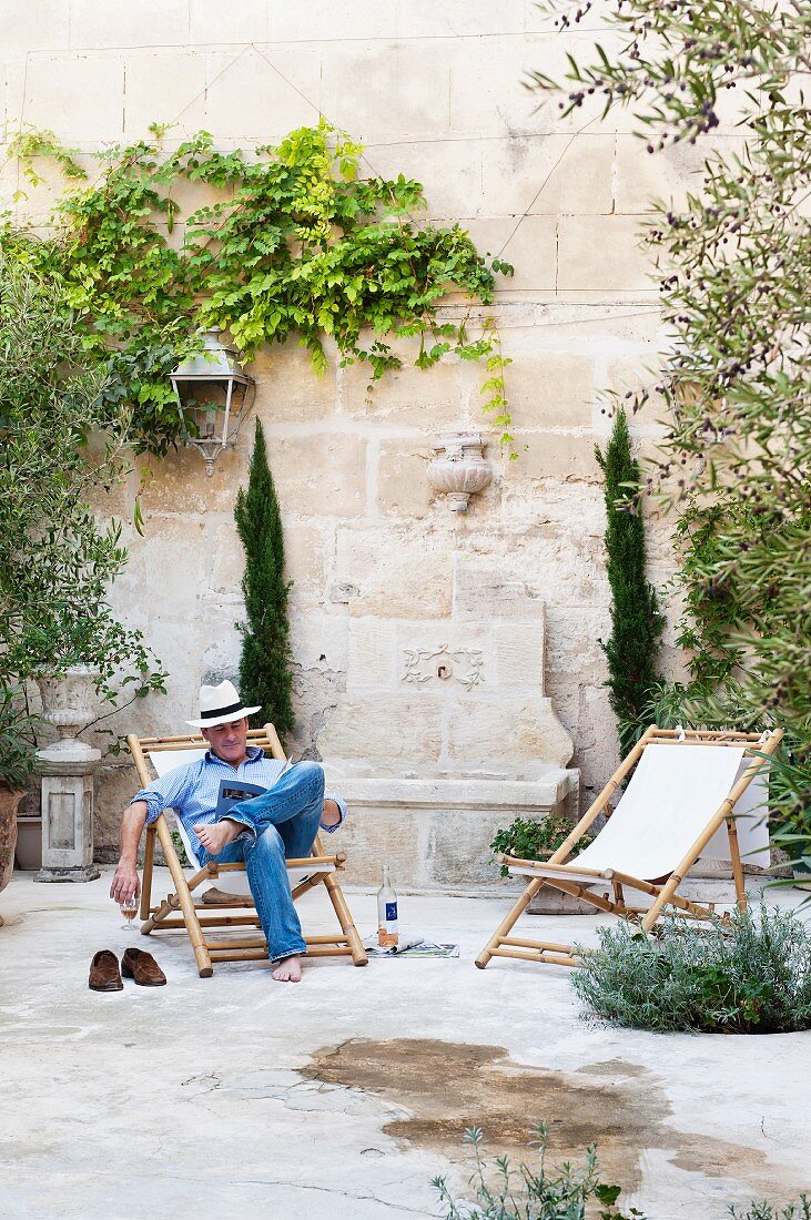 Man reading in wooden deckchair in summery courtyard with high, climber-covered wall of Provençal townhouse