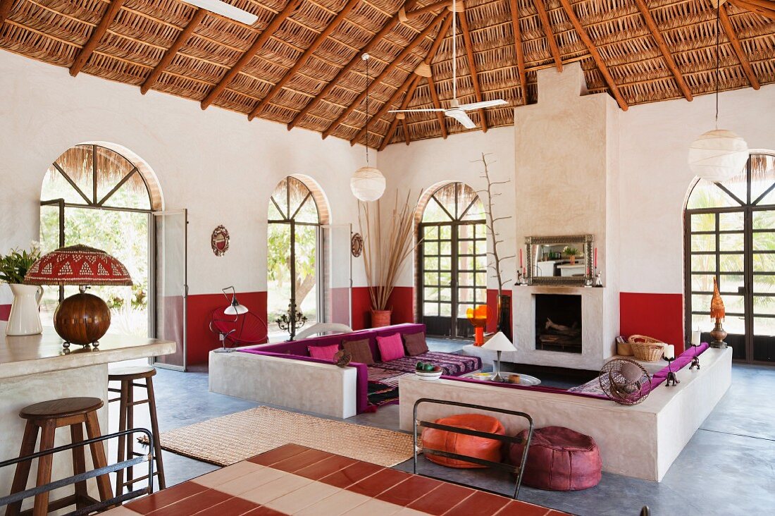 Offener Wohnraum mit Palmendach über gemauerte Sitzbänke und farbige Polsterelemente vor Rundbogentüren