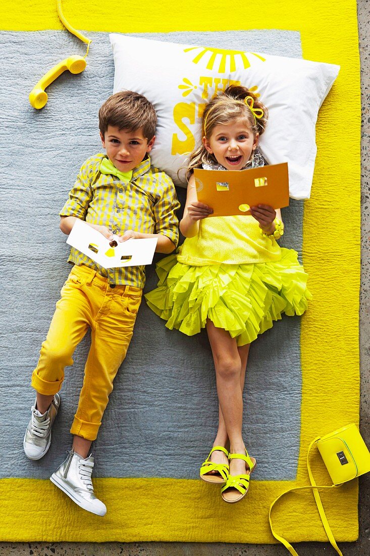 Grey rug with yellow border; little girl and boy wearing shades of yellow lying on rug