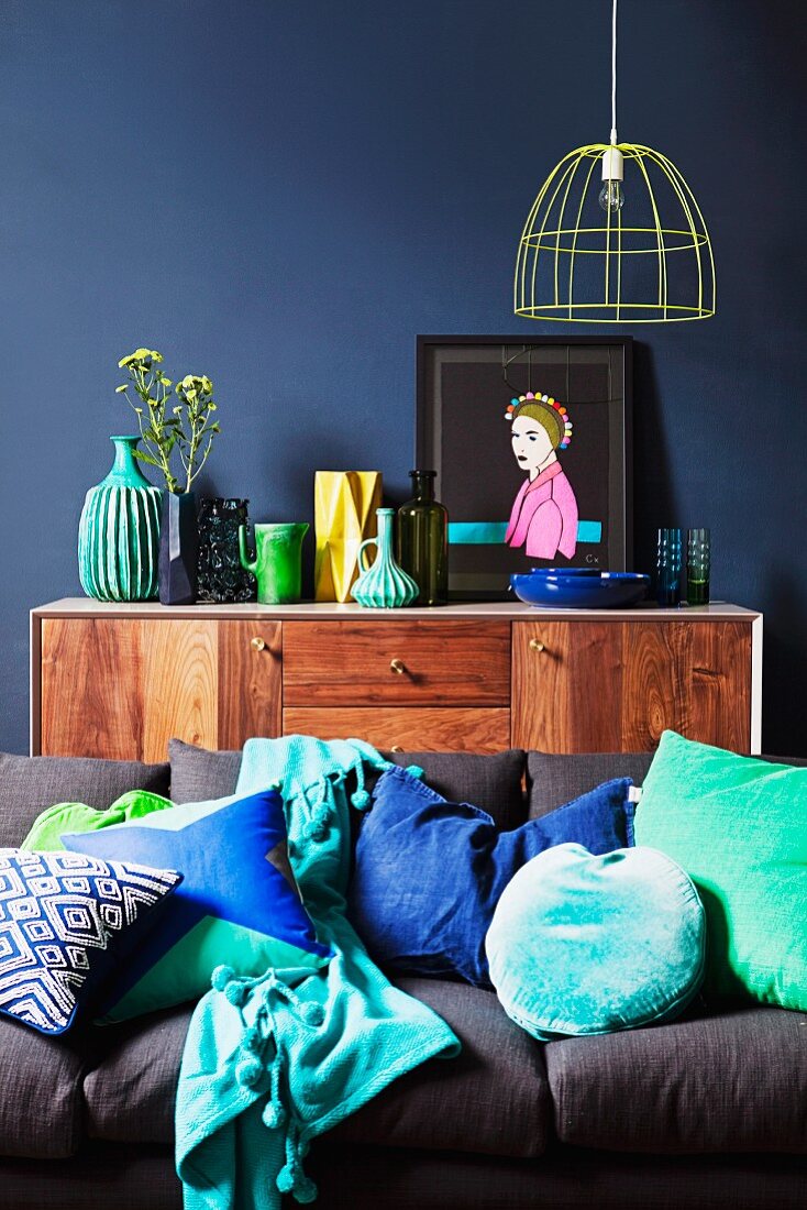 Sideboard im Retrolook vor blauer Wand; im Vordergrund eine graue Couch mit grünen und blauen Kissen