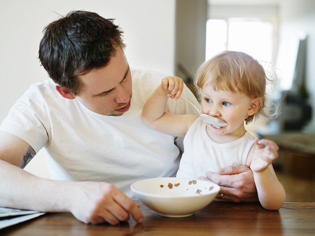 Kleines Mädchen auf dem Schoss des Vaters isst Müsli zum Frühstück