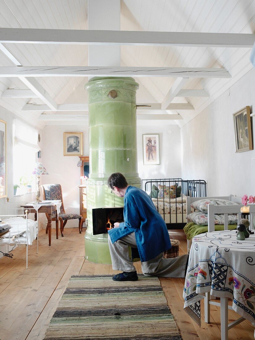 Mehrzweckraum in renoviertem Landhaus mit zentralem Kachelofen unter weiss lackiertem, offenem Dachstuhl; Frau beim Anzünden des Ofens