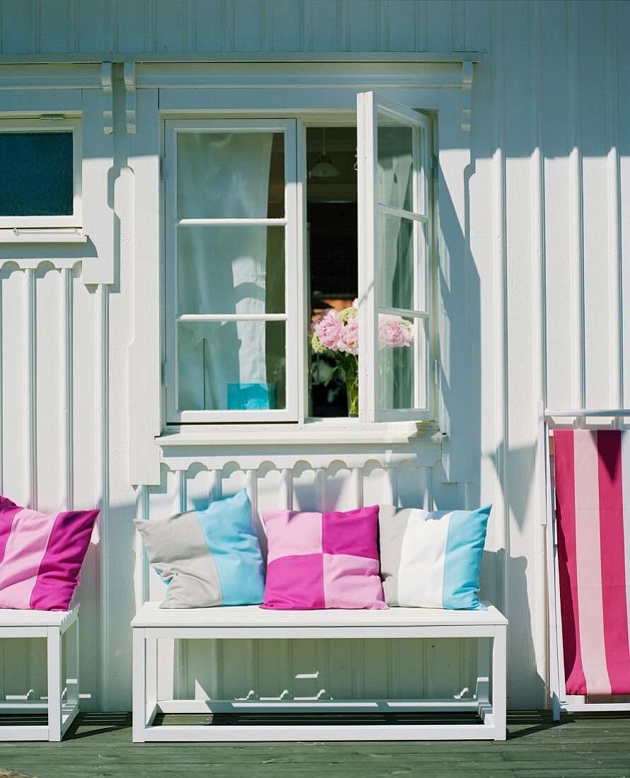 Bunte Kissen auf Sitzbank vor weiss gestrichenem Holzhaus an einem sonnigen Tag
