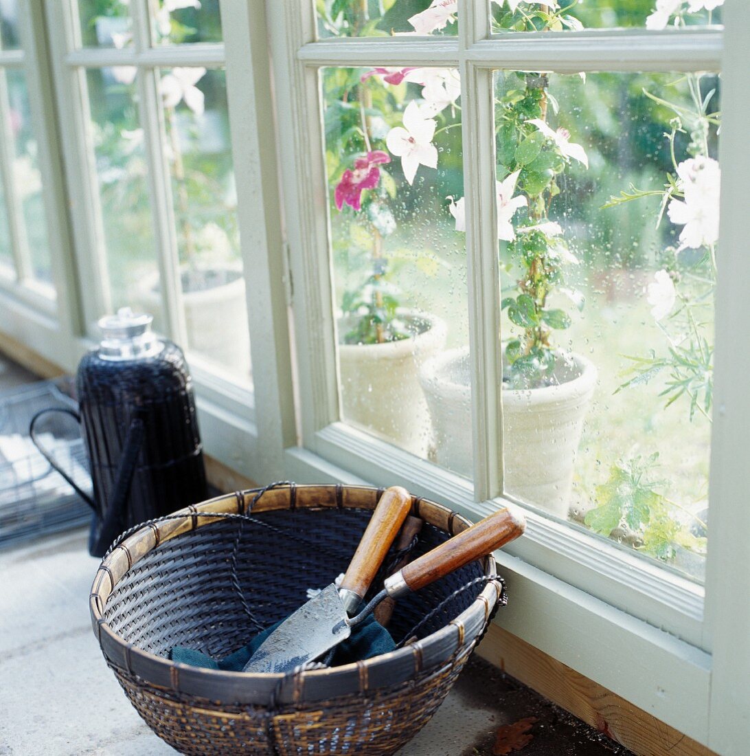 Gardening tools in basket on floor next to French window with view of potted plants outside