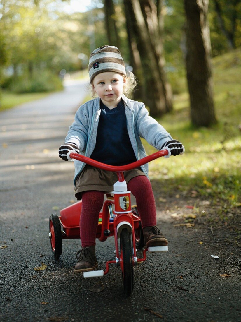 Blonde little girl wearing striped hat and gloves riding a tricycle along a woodland path