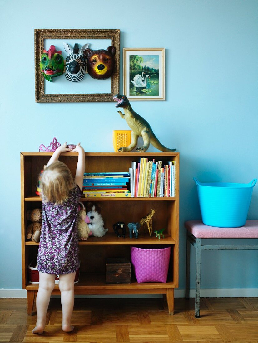 Kleines Mädchen vor Regal mit Spielzeug; Tiermasken in Bilderrahmen und Dinosaurierfigur vor hellblau getönter Wand