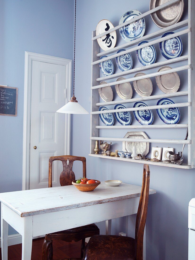 Blue and white crockery and pewter plates in plate rack on lavender-painted kitchen wall; old dining table and antique wooden chairs