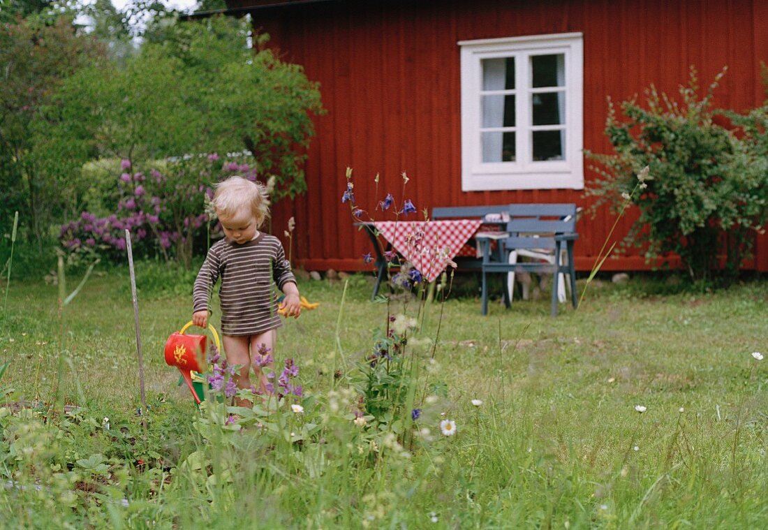 Kind mit nackten Beinen beim Blumengiessen vor rotem, schwedischem Ferienhaus