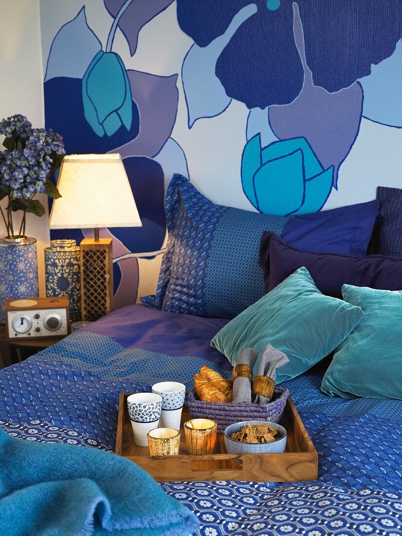 Breakfast tray on double bed in front of decorative, floral wall paper and textiles in many nuances of blue