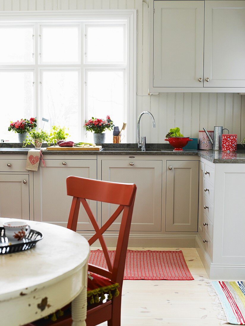 Vases of flowers on window sill in white, fitted kitchen; wooden chair at old, round dining table in foreground