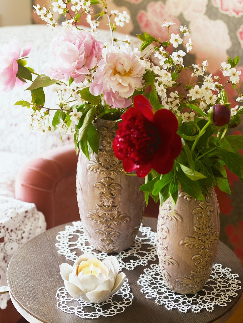 Peonies in two ornate vases and flower-shaped candle on doilies