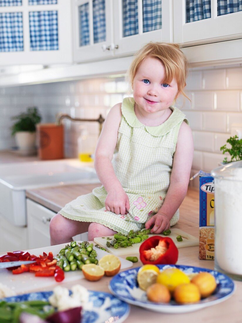 Kleines Mädchen sitzt auf Küchenarbeitsplatte und schneidet Gemüsestücke