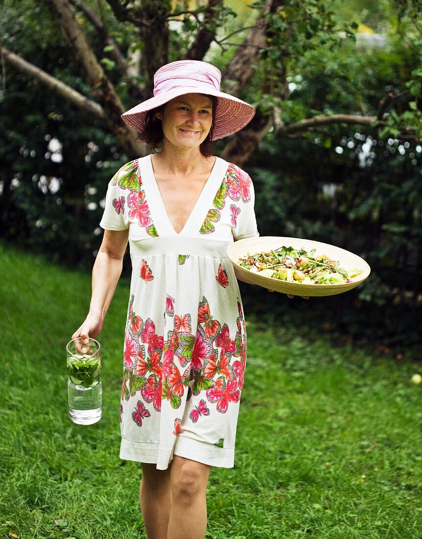 Frau in Sommerkleid trägt Salat & Gtränkekrug in den Garten