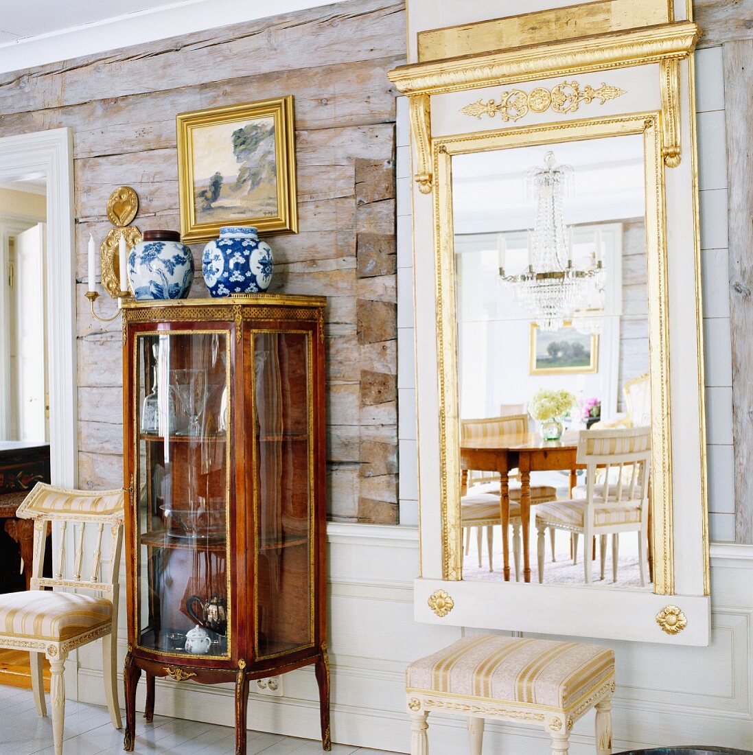 Antike Glasvitrine und vergoldeter Spiegel vor den rustikalen Wänden eines Blockhauses