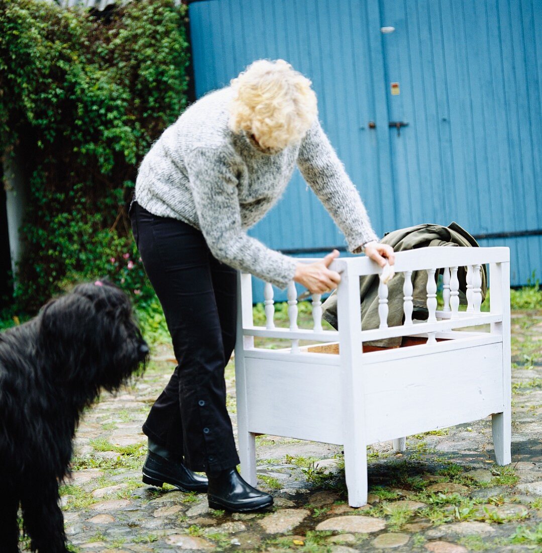 Frau mit schwarzem Hund in gepflastertem Eingangshof beim Ausbessern einer weissen Sitzbank