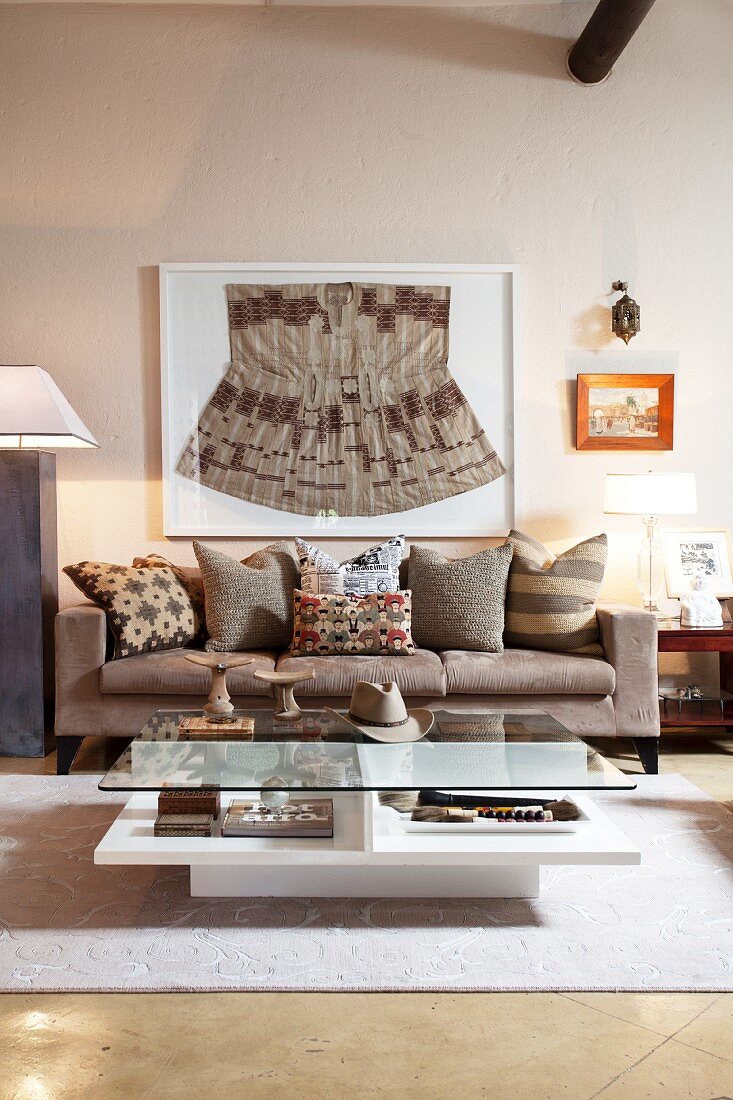Ethno-Sammelstück hinter Glas über grauer Couch mit streng geordneten Zierkissen im Ethnolook in modernem Wohnzimmer; auf dem Couchtisch Sammelstücke aus Holz