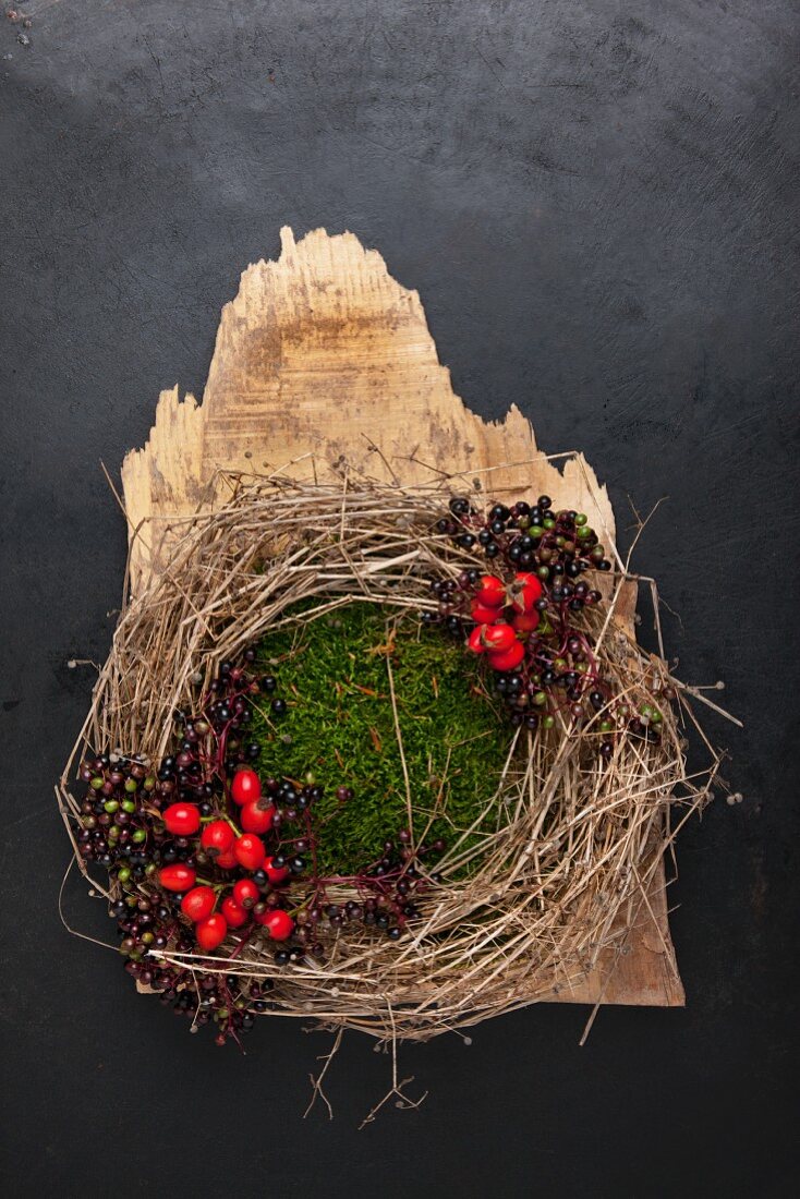 Small wreath of foraged natural items; fragment of wood, moss, twisted dried grasses, elderberries and rosehips on dark metal surface