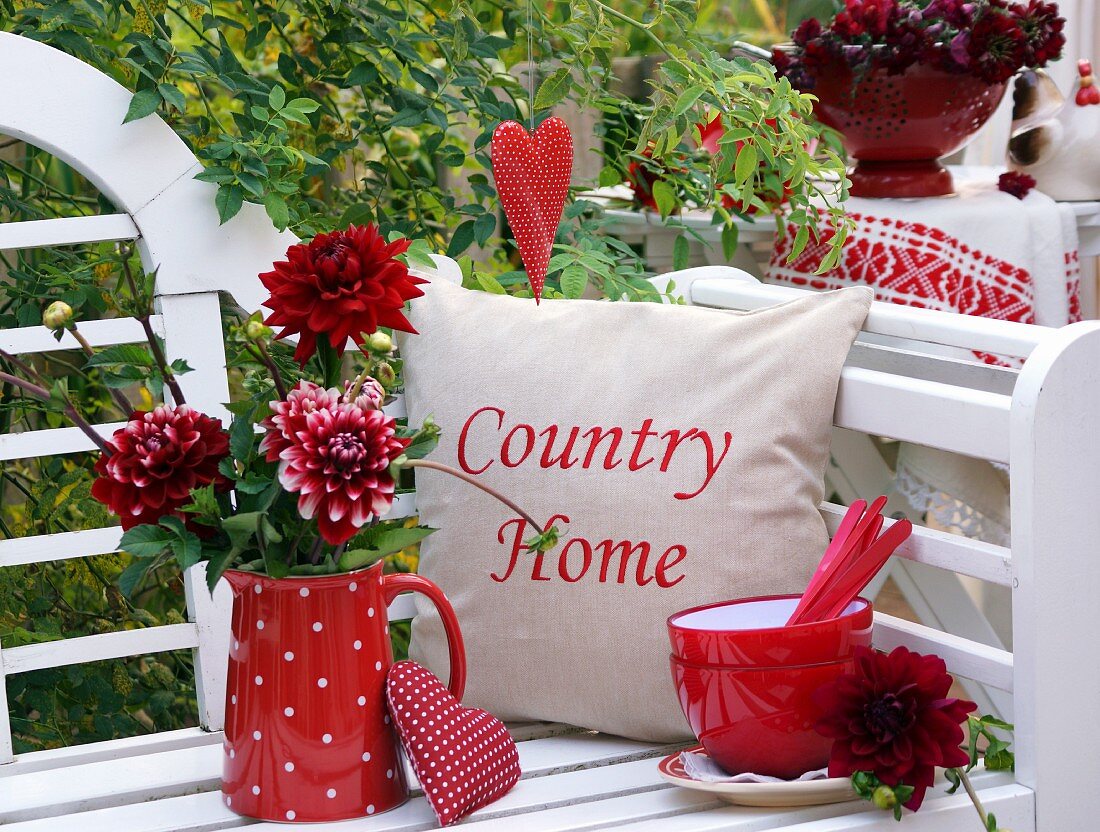 Red dahlias in red, polka-dotted jug next to stack of bowls and cushions on white garden bench