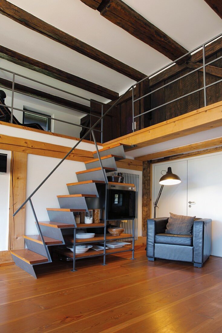 Metalltreppe mit Holz Trittstufen und integriertem Regal neben Stehleuchte und Sessel unter Galerie in rustikalem Wohnraum
