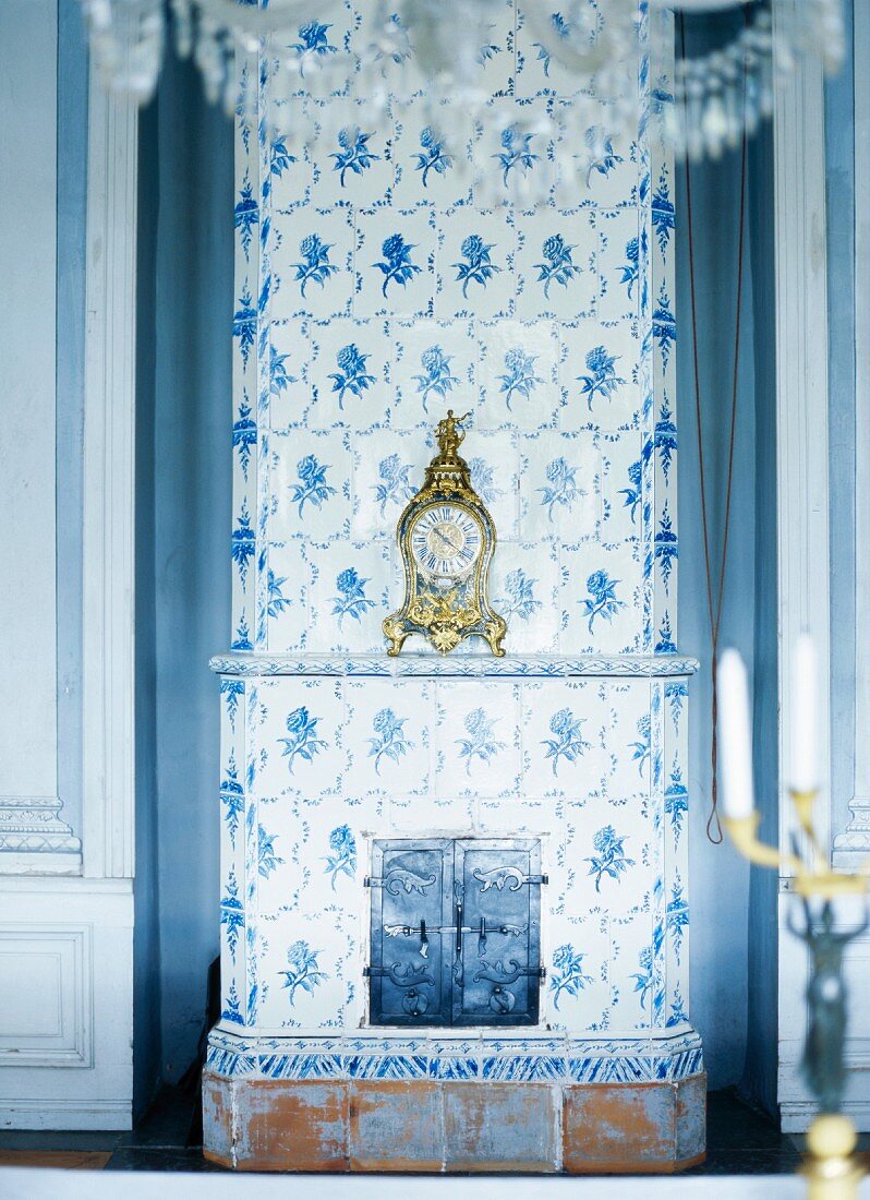 Old fireplace with white and blue ceramic tiles and gilt mantel clock on mantelpiece